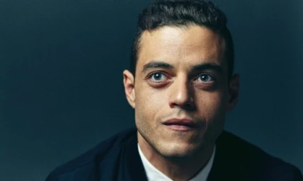 Close-up portrait of actor Rami Malek looking upward with a serious expression against a dark background