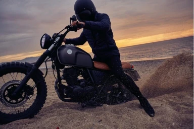 A rider in a black helmet and quilted jacket on a motorcycle stirring up sand on a beach at sunset