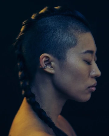 Profile portrait of a person with a shaved head and a long central braid against a dark background