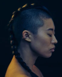 Profile portrait of a person with a shaved head and a long central braid against a dark background