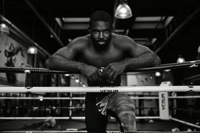 Black male boxer leaning on the ropes of a boxing ring inside a gym, captured in monochrome by Coty Tarr.