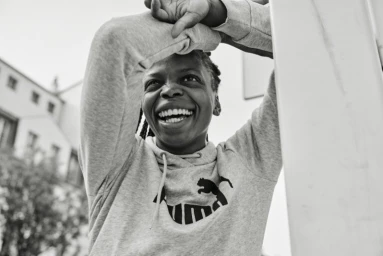 Smiling Black woman in a grey hoodie with arms raised and hands clasped behind her head, photographed by Coty Tarr.
