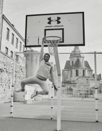 Black man dunking a basketball, hanging from a hoop on an outdoor court