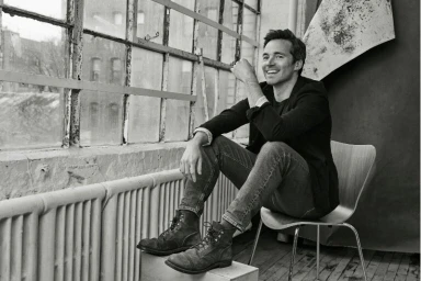 Monochrome portrait of a smiling man in a dark jacket and jeans seated on a chair next to large industrial factory windows