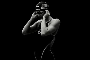 Black and white portrait of a female swimmer adjusting her goggles and swim cap against a dark background.