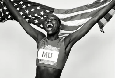 Black female athlete celebrating with arms raised holding an American flag in a black and white portrait by Coty Tarr