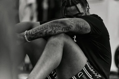 Black and white portrait of a black man seated with arms resting on his knees, showing extensive arm tattoos