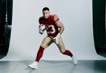 Football player in red jersey and gold pants holding a football against white backdrop