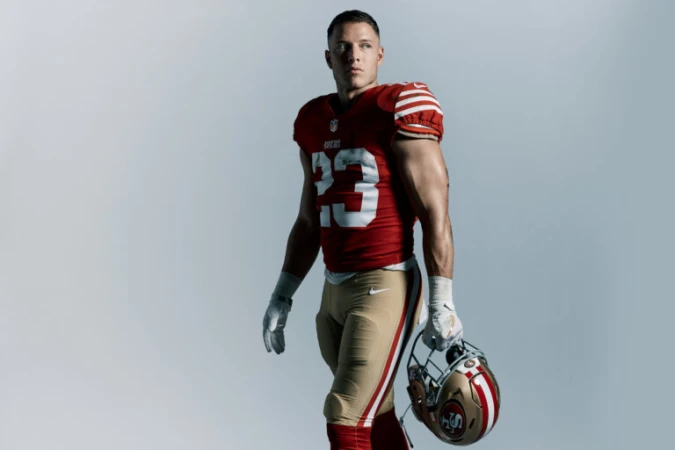 Christian McCaffrey in a red and gold football uniform holding a helmet against a light grey background