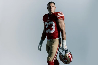 Christian McCaffrey in a red and gold football uniform holding a helmet against a light grey background