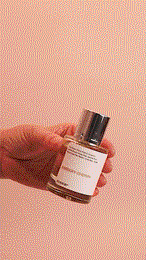 Hand holding a bottle of perfume with a white label against a light pink background in an indoor setting, beauty shot.