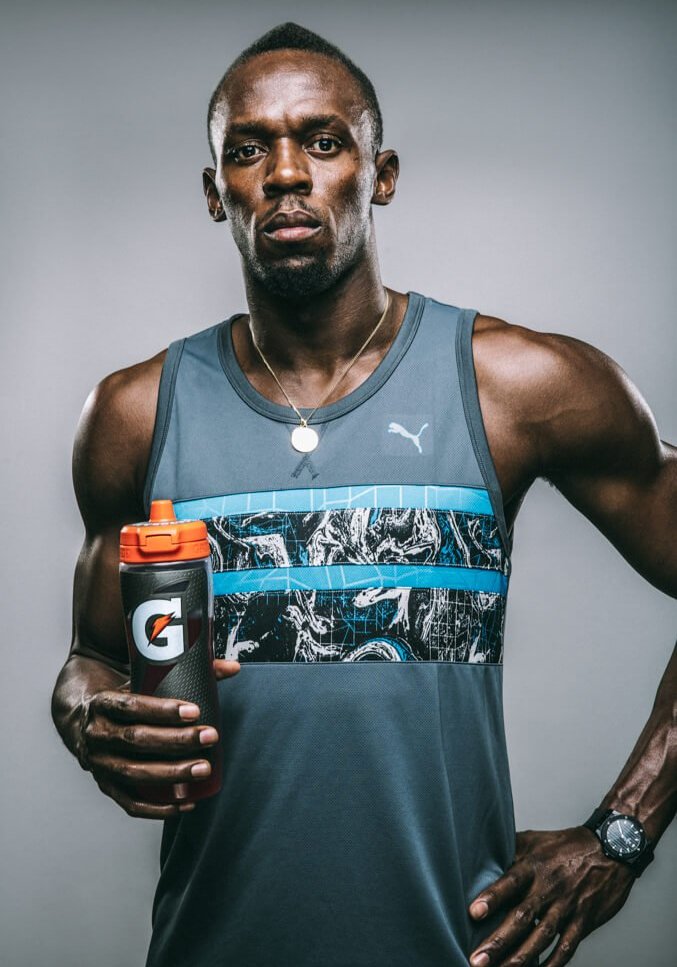 Adult male athlete Usain Bolt poses in a tank top holding a Gatorade bottle in a medium shot against a neutral grey background.