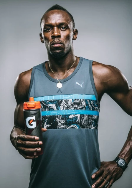 Adult male athlete Usain Bolt poses in a tank top holding a Gatorade bottle in a medium shot against a neutral grey background.