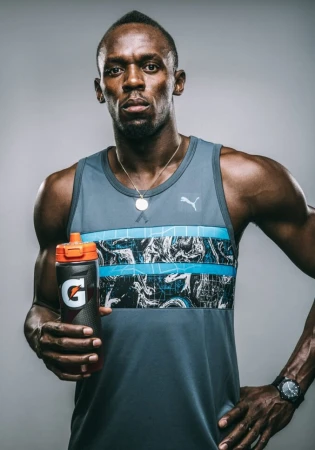 Adult male athlete Usain Bolt poses in a tank top holding a Gatorade bottle in a medium shot against a neutral grey background.