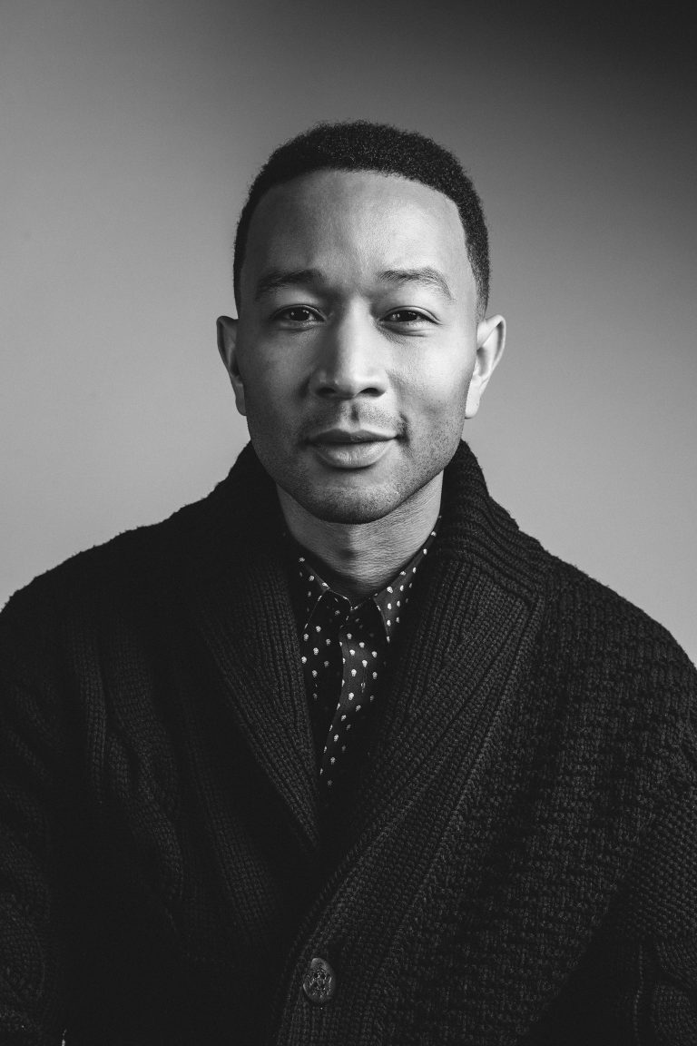 Black and white portrait of a man wearing a cardigan and collared shirt, gazing at the camera against a plain gray background.