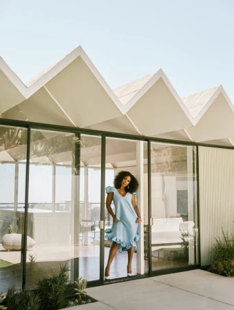 A striking full shot captures a smiling woman in a stylish blue dress, standing in a doorway with a modern home and outdoor patio.
