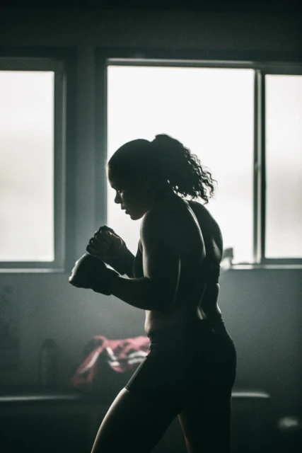 Silhouette of female boxer in sportswear and boxing gloves in training stance near windows in a gym setting.
