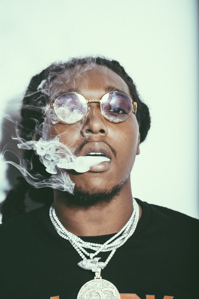 Portrait of a man with dreadlocks wearing round sunglasses and several necklaces, exhaling a large cloud of smoke on a white background.