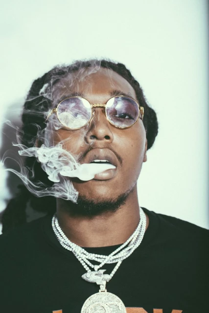 Portrait of a man with dreadlocks wearing round sunglasses and several necklaces, exhaling a large cloud of smoke on a white background.