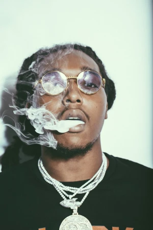 Portrait of a man with dreadlocks wearing round sunglasses and several necklaces, exhaling a large cloud of smoke on a white background.