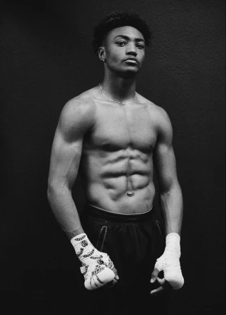 Striking portrait of an African American man with a muscular physique, bare-chested and wrapped hands, set against a stark black background.