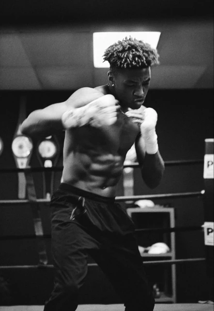 Monochrome shot of a muscular man in boxing stance with hand wraps, focused expression, set in an indoor training facility.