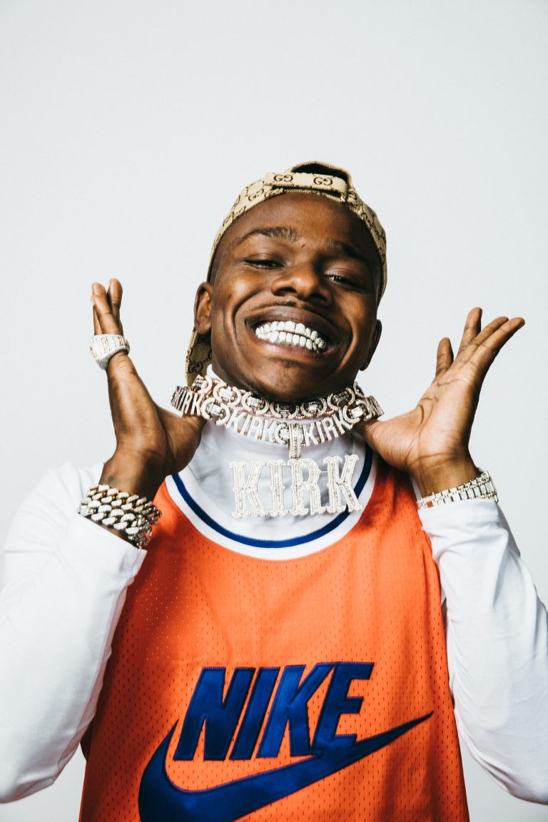 Smiling adult male wearing a cap and orange jersey with silver chains and a ring against a bright background in a portrait shot.