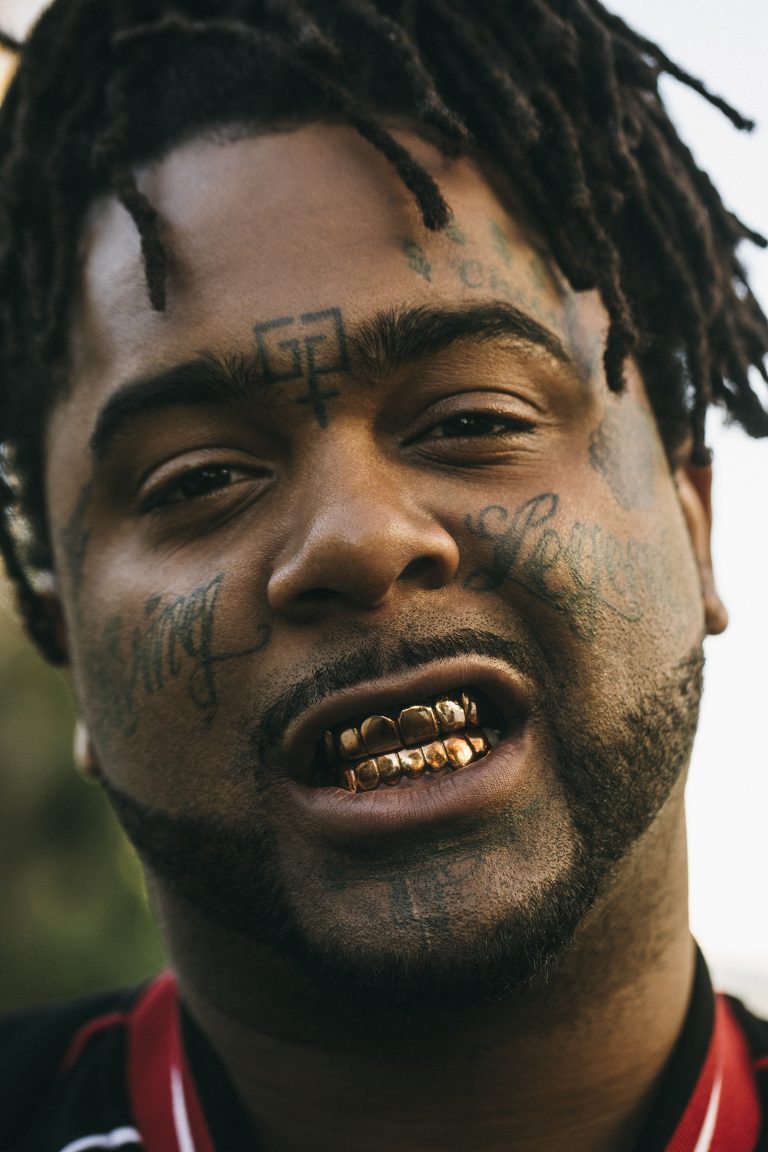 Portrait of a man with dreadlocks, face tattoos, and gold teeth. The image focuses on the man's face, and upper torso.