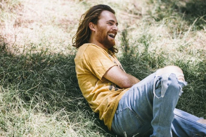 Adult male with long brown hair, wearing a yellow t-shirt and ripped jeans, is sitting in green grass with a happy facial expression.