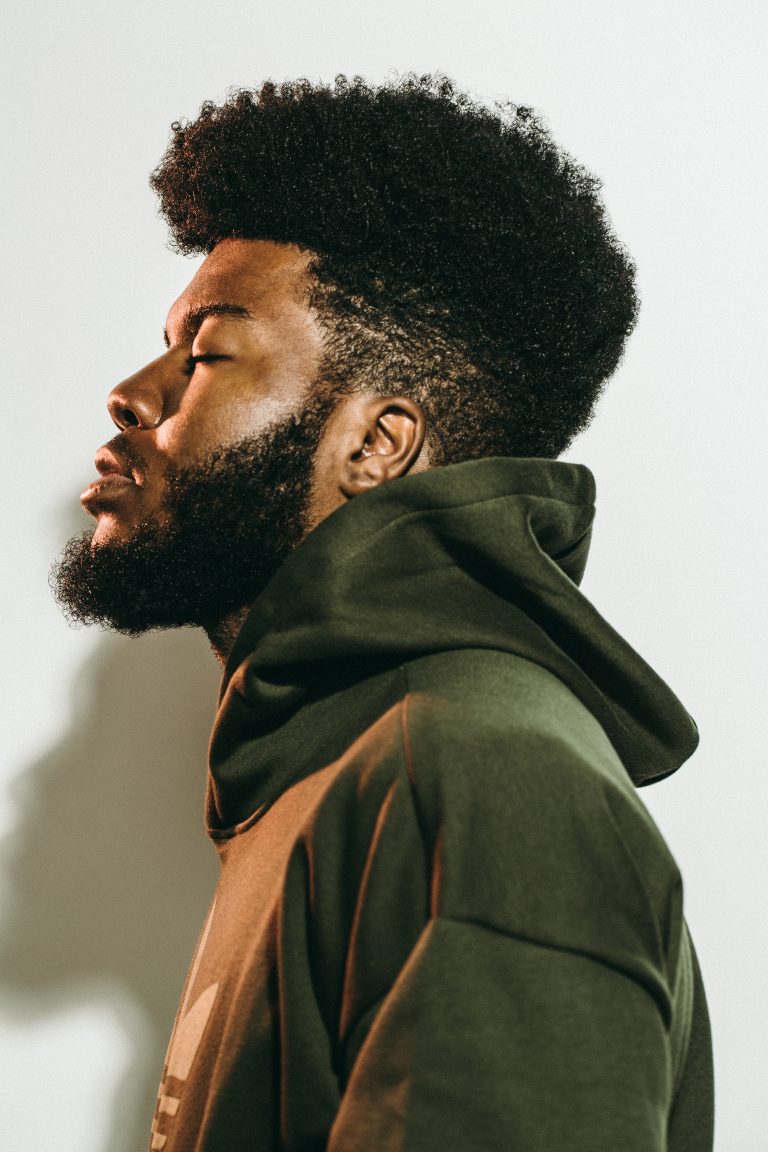 Profile portrait of a Black man with a prominent afro and beard wearing an olive green hoodie against a white background