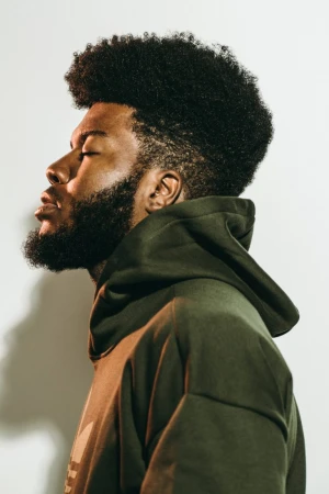 Profile portrait of a Black man with a prominent afro and beard wearing an olive green hoodie against a white background