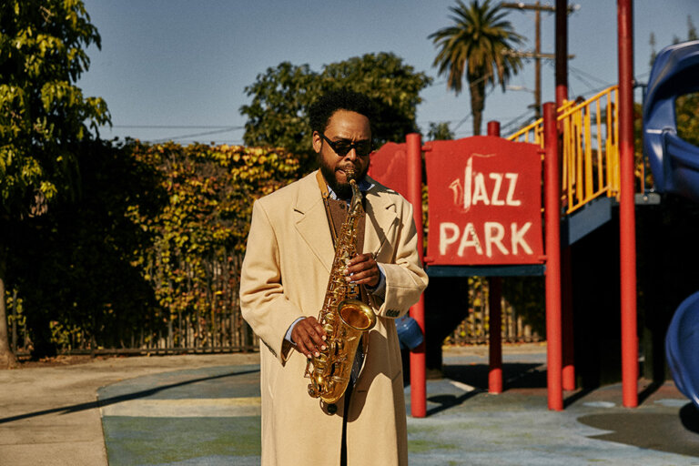 Adult male playing a saxophone in a park with playground equipment, wearing a tan coat, sunglasses, and a blue shirt on a sunny day.