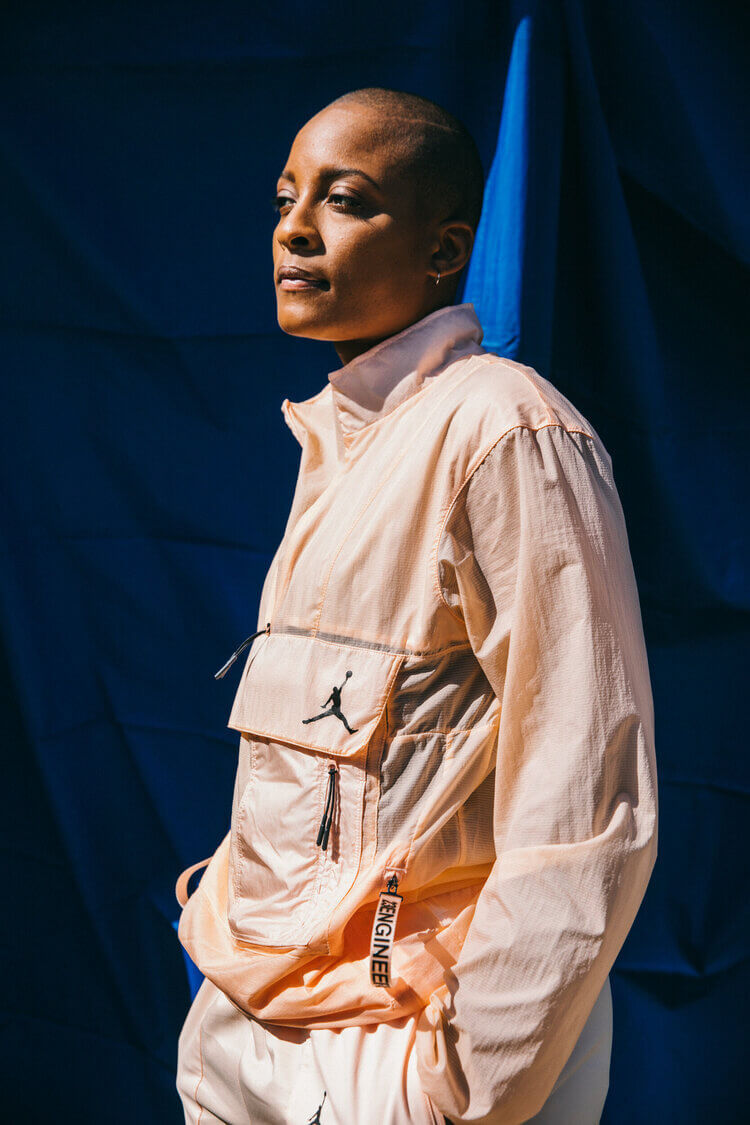 A woman in a light peach-colored jacket stands against a dark blue background. The jacket features a visible Air Jordan logo.