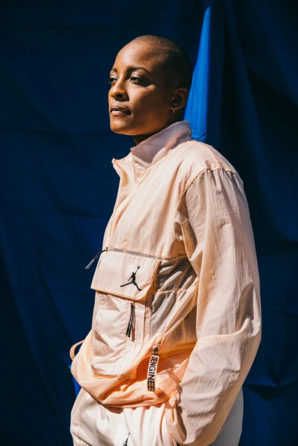 A woman in a light peach-colored jacket stands against a dark blue background. The jacket features a visible Air Jordan logo.