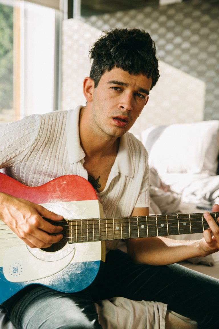Adult male playing a red, white, and blue acoustic guitar indoors, wearing a short-sleeved polo shirt and jeans.