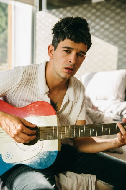 Adult male playing a red, white, and blue acoustic guitar indoors, wearing a short-sleeved polo shirt and jeans.