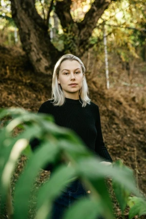 Portrait of a blonde woman wearing a black turtleneck shirt standing outdoors with a large tree and foliage in the background.