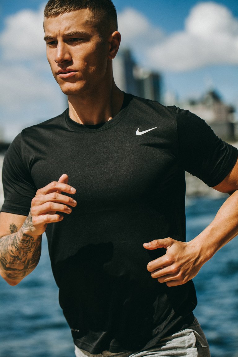 Athletic man in black Nike t-shirt running outdoors with city skyline, water in the background, capturing a fitness and active lifestyle scene.
