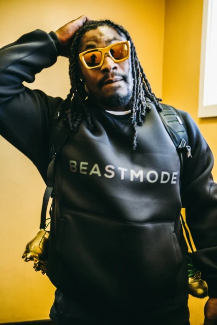 An African-American man wearing a black hoodie and yellow sunglasses poses indoors. He is also wearing a backpack. The sweatshirt has 'BEASTMODE' on it.