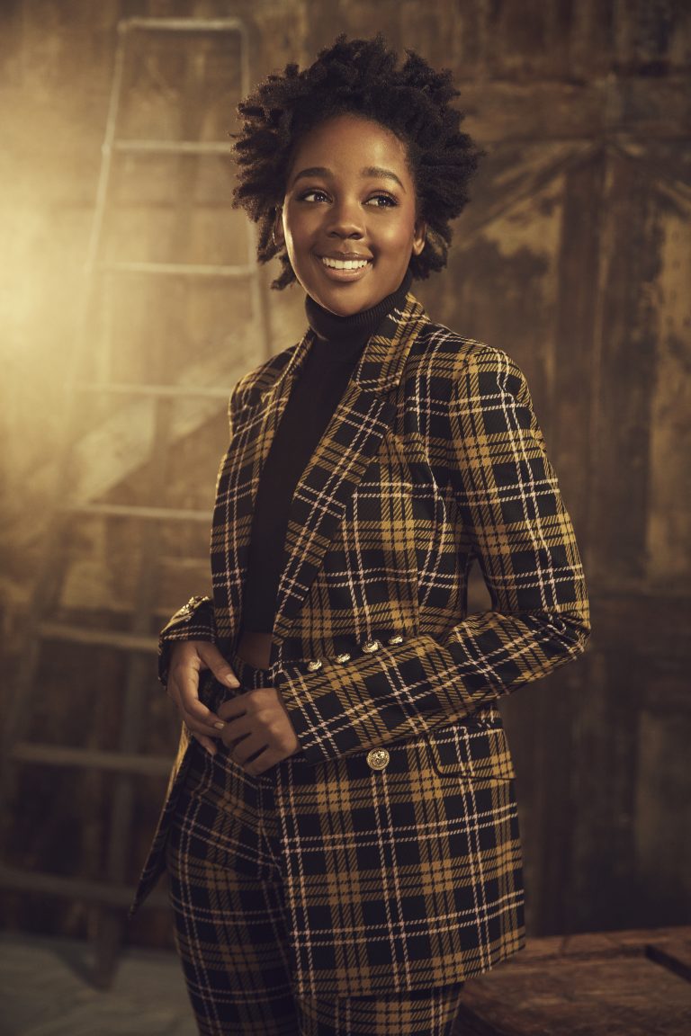 A smiling black woman with natural hair wears a black turtleneck and a yellow plaid suit in a portrait with a ladder background.