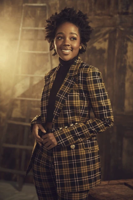A smiling black woman with natural hair wears a black turtleneck and a yellow plaid suit in a portrait with a ladder background.