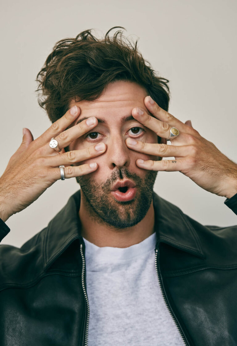Close-up portrait of a man in a black leather jacket making a playful face, covering his eyes with his fingers, against a neutral backdrop.