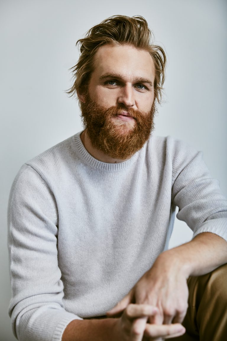 Portrait of a man with a beard wearing a light grey sweater. The man has light brown hair and is sitting indoors with a neutral expression.
