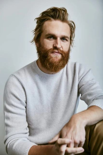Portrait of a man with a beard wearing a light grey sweater. The man has light brown hair and is sitting indoors with a neutral expression.