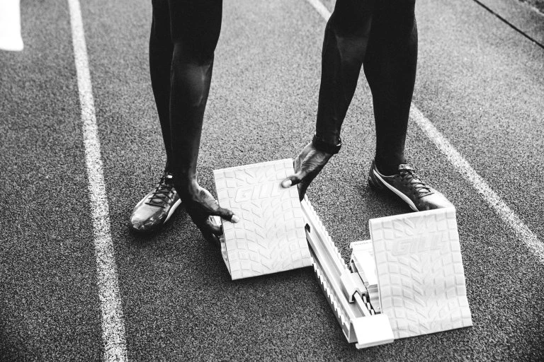 Close-up of an athlete at the starting line, placing hands on starting blocks on a textured black running track.