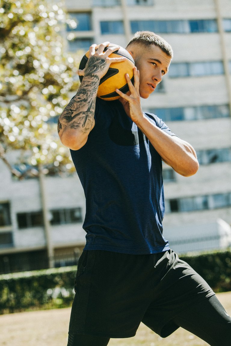 Young man holding a basketball against his head with tattooed arms, wearing a navy t-shirt and black shorts outdoors
