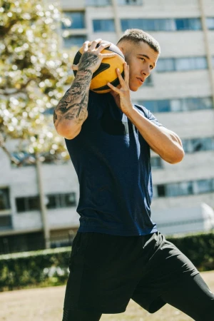 Young man holding a basketball against his head with tattooed arms, wearing a navy t-shirt and black shorts outdoors