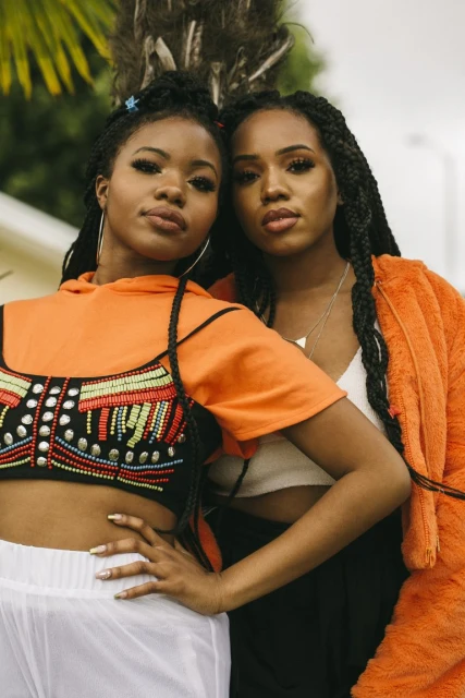 Two young Black women pose outdoors in colorful casual fashion. One wears an embellished crop top and the other an orange hoodie.