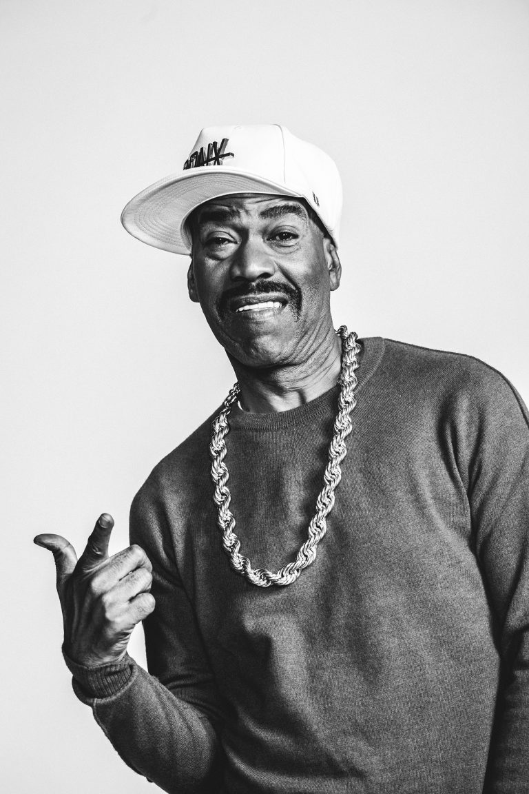 Black man wearing a baseball cap and thick chain necklace pointing forward in a studio portrait by G L Askew Ii