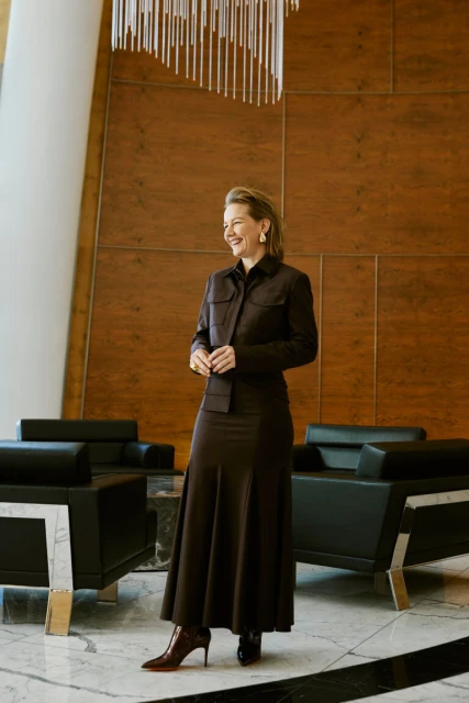 Elegant woman in a brown skirt suit smiles in a modern lobby with marble floors, black armchairs, wood paneled walls, and a chandelier.